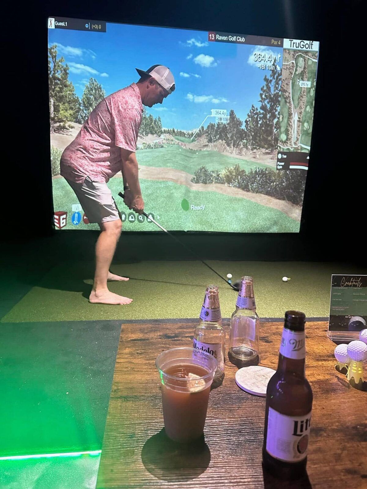A golfer lining up a tee shot on the simulator at Grip It Virtual Golf with drinks on the table in front